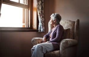 An elderly woman looking at the window sitting in her bedroom.
