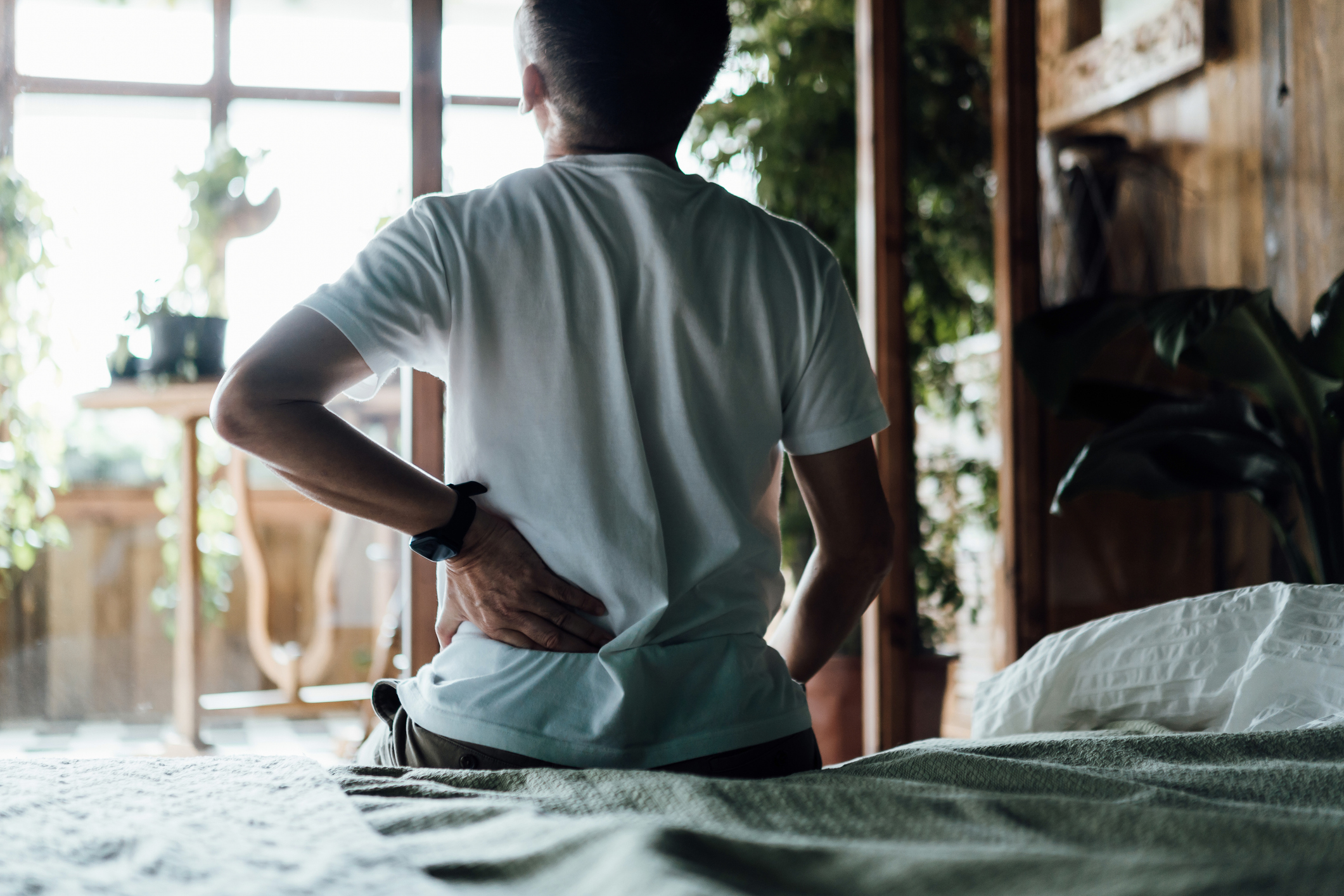 From behind, an elderly man massages his sore back muscles as he sits on the bed, dealing with back pain.