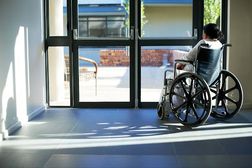 A woman in a wheelchair sits by the front door of a nursing home and looks out the window.