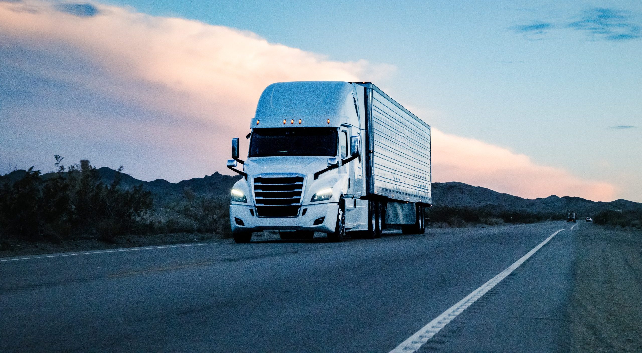 A white semi-truck driving on an open highway at dusk with mountains in the background.