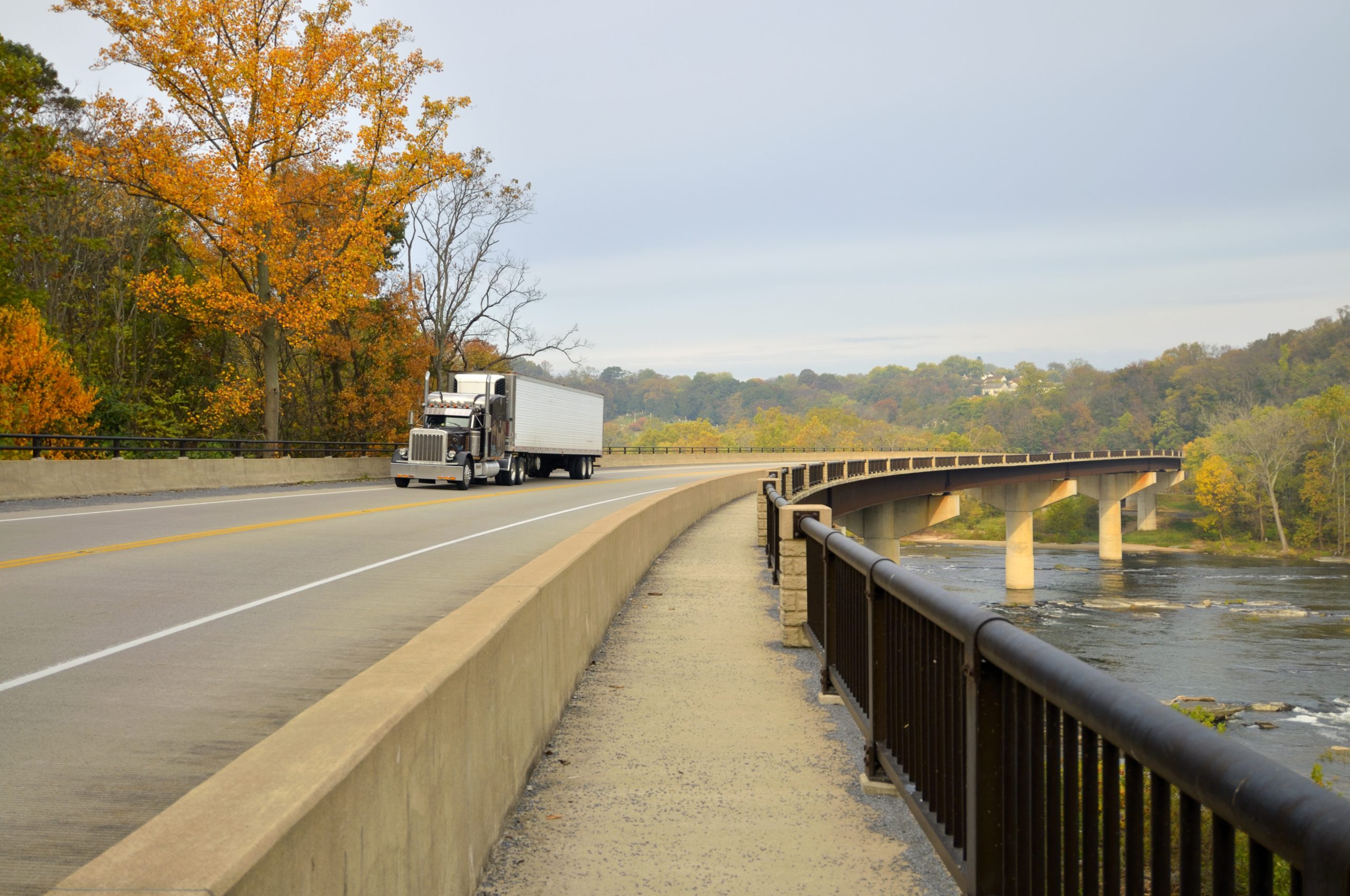 A semi-truck driving across a bridge surrounded by autumn trees and a river below.